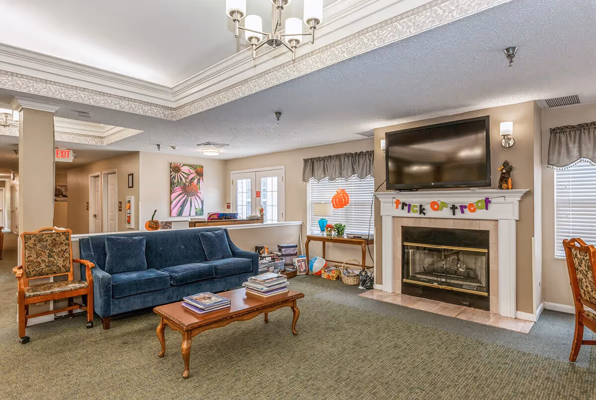 A cozy living room area in a senior living facility with a blue sofa, wooden coffee table with books, and a fireplace with a mounted TV above it. The fireplace mantel is decorated with a colorful 'trick or treat' banner and small Halloween decorations. There are windows with blinds and valances, a side table with a lamp and plants, and a hallway leading to other rooms.