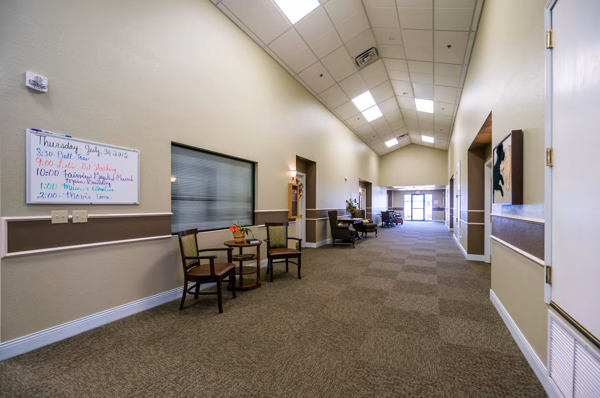 A long, carpeted hallway in a senior living facility with high ceilings and recessed lighting. Along the walls are small seating areas with chairs and tables, some decorated with plants and flowers. A whiteboard on the left wall displays a schedule of activities for Thursday, July 30, 2015. The hallway leads to a glass exit door at the far end.