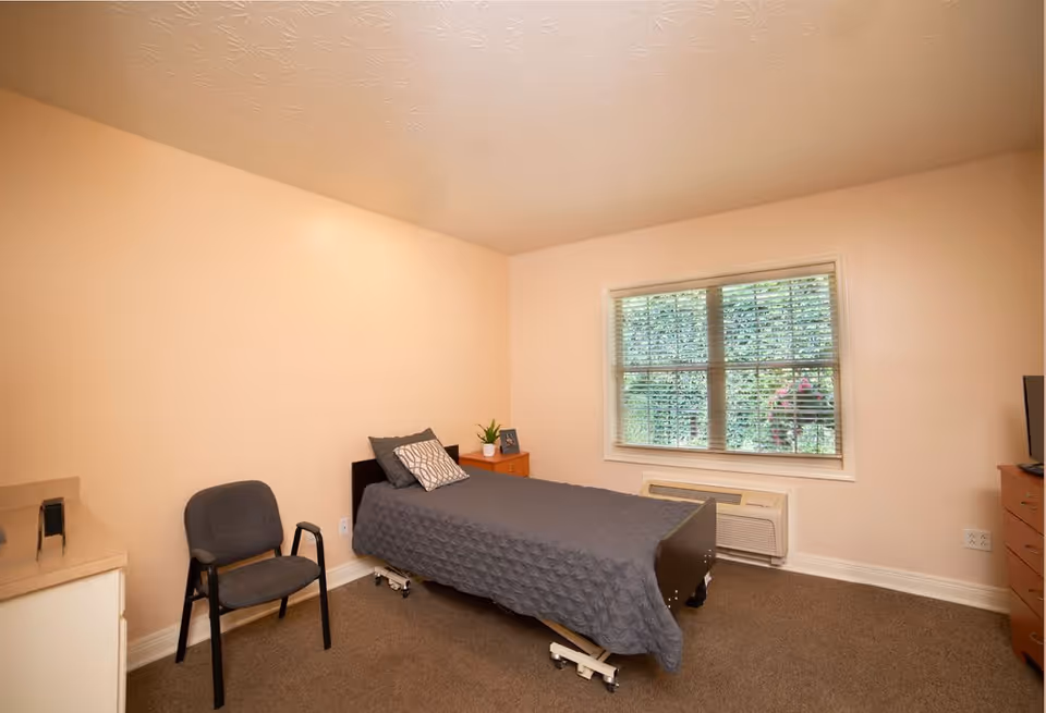 A simple bedroom with a single bed covered with a gray quilt and a patterned pillow. Next to the bed is a small wooden nightstand with a small plant and a framed photo. There is a gray chair with armrests near the bed. A window with blinds shows greenery outside. The room has beige walls and carpeted floor.