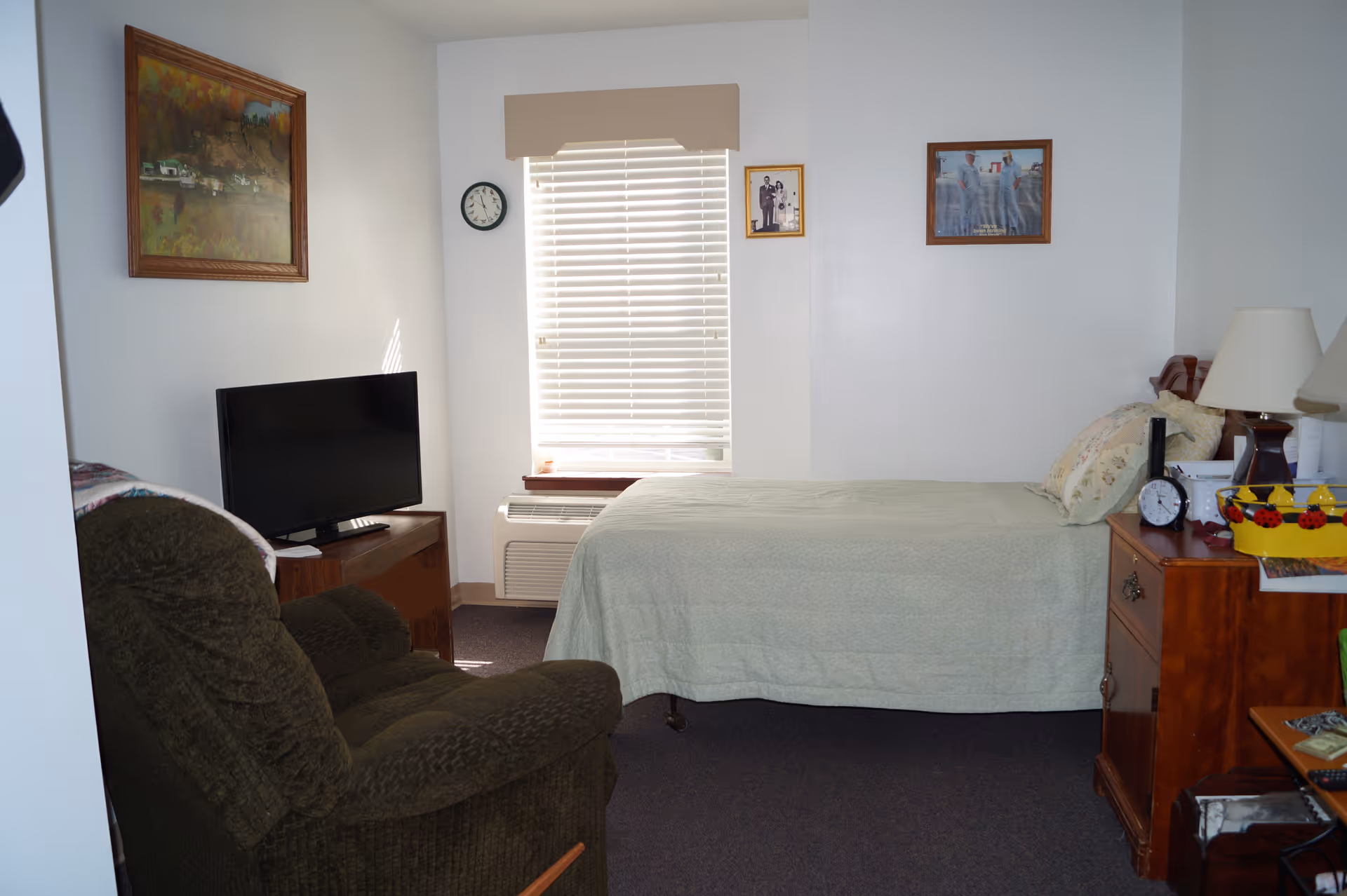 A small furnished bedroom with a single bed, armchair, television, and wooden nightstand beneath a window with blinds.