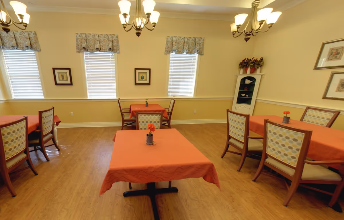 A dining room with several tables covered in orange tablecloths, each with a small flower centerpiece. The room has wooden flooring, light yellow walls, three windows with blinds and patterned valances, framed artwork on the walls, and two chandeliers hanging from the ceiling. There is also a white corner shelf with decorative items and plants.