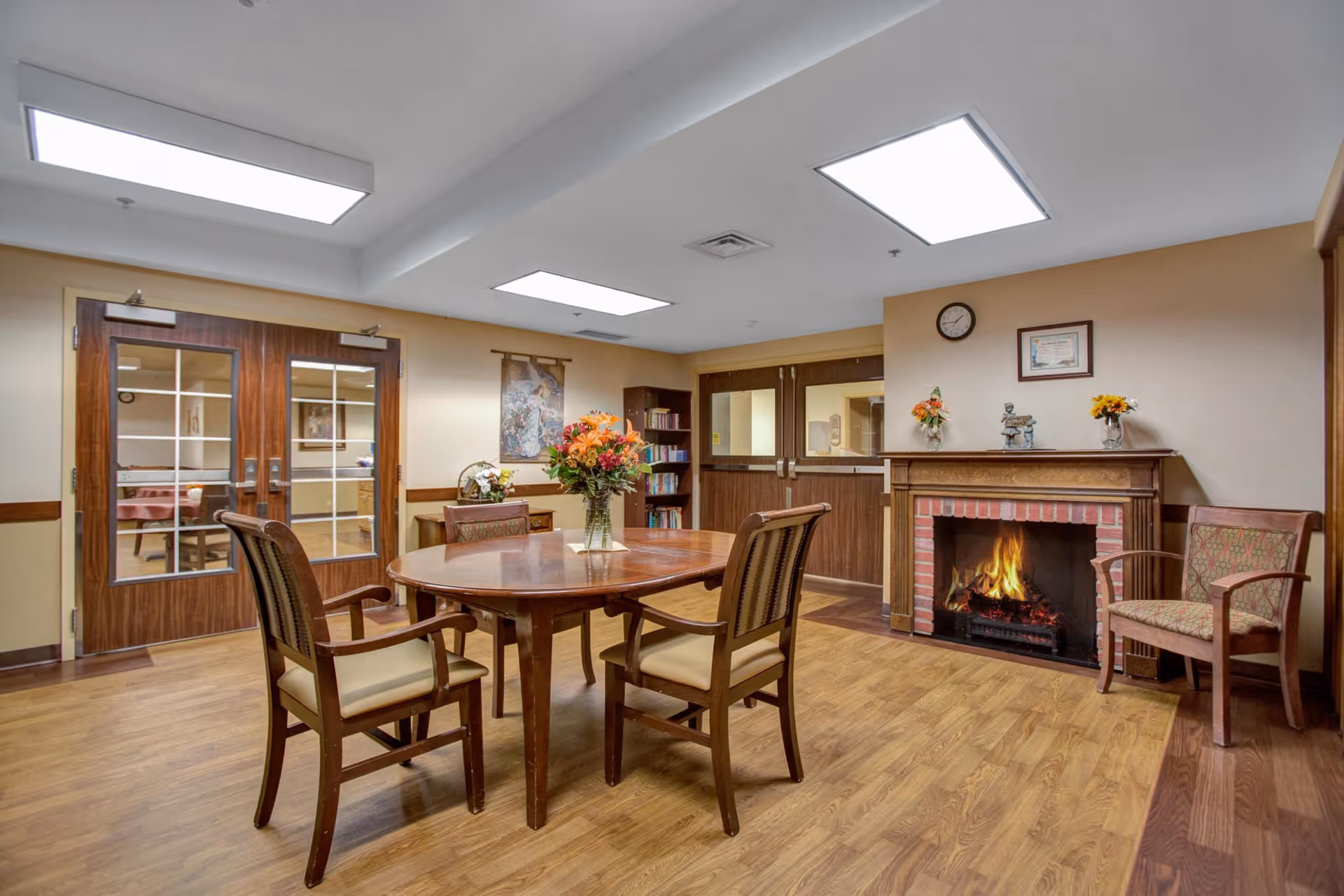 Communal dining area with a round wooden table and chairs, a lit fireplace, bookshelves, and double doors.