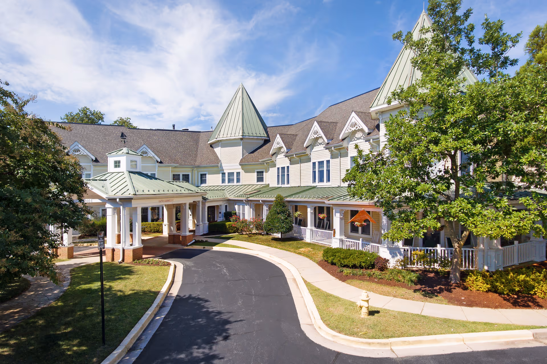 Front entrance of a large Victorian-style senior living facility with a covered porte-cochère, green metal roofs, driveway and landscaped grounds.