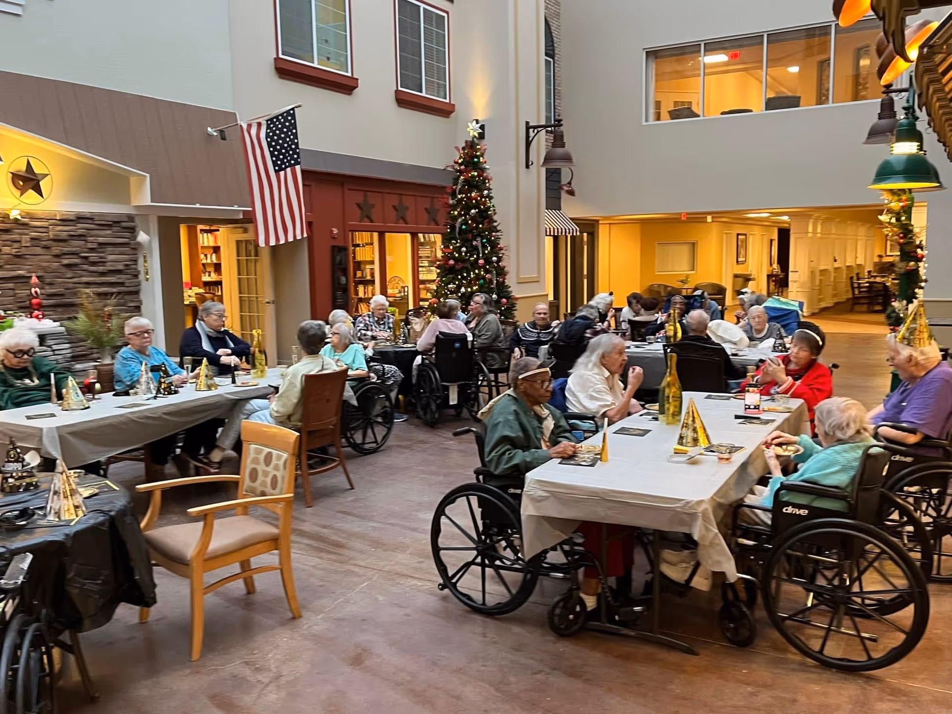 A group of elderly residents, many in wheelchairs, seated around tables in a decorated indoor common area with a Christmas tree and American flag visible. The room has warm lighting and festive party hats on the tables, suggesting a holiday celebration.