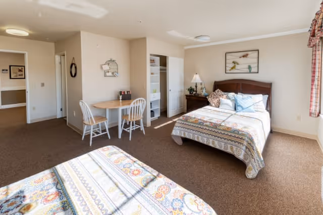 A bright and spacious bedroom in a memory care facility featuring two beds with patterned quilts, a small round wooden table with two chairs, a nightstand with a lamp and plant, a closet with open shelving, and a window with plaid curtains allowing natural light to fill the room.