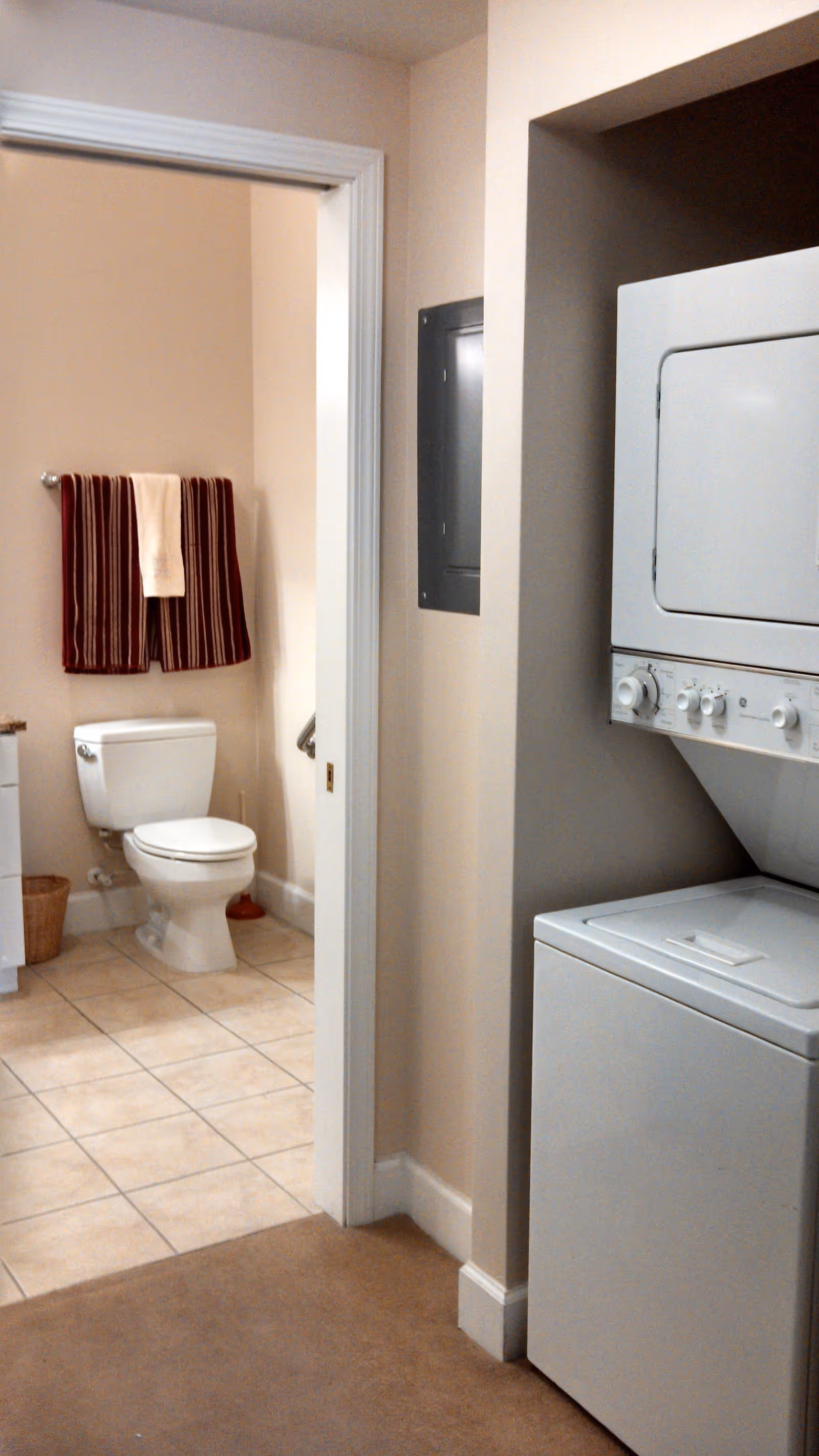 Stacked washer/dryer in a hallway next to an open bathroom doorway showing a toilet and towels on a rack.