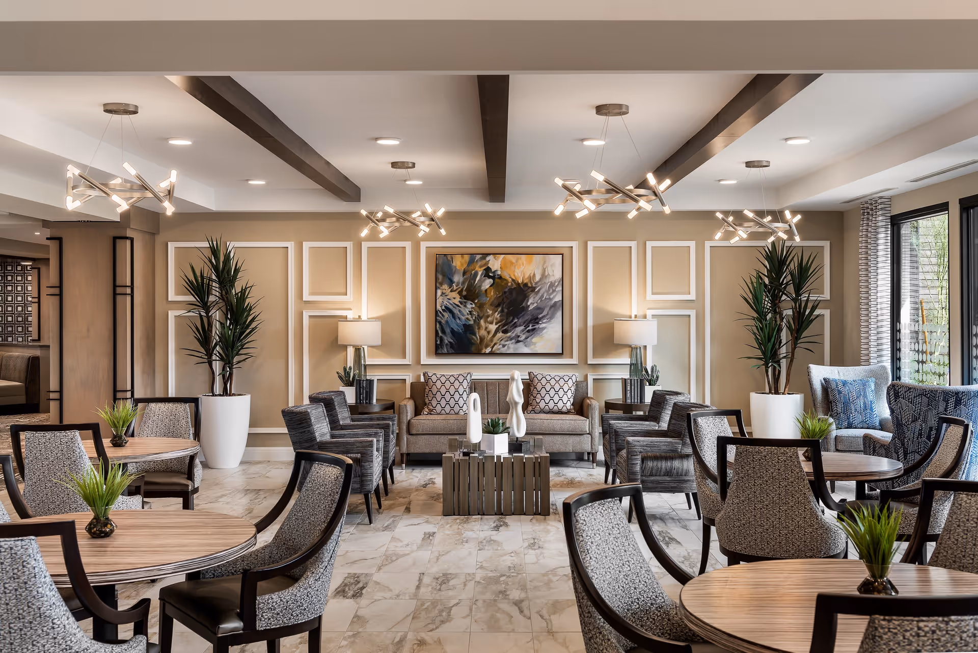 A spacious and elegant common area with multiple round wooden tables surrounded by patterned chairs. The room features a beige wall with decorative white molding, two large potted plants, modern light fixtures hanging from the ceiling, and a sofa with patterned pillows centered against the wall. There are also two side tables with lamps and decorative sculptures on a coffee table in front of the sofa. Large windows with curtains allow natural light to enter the room.