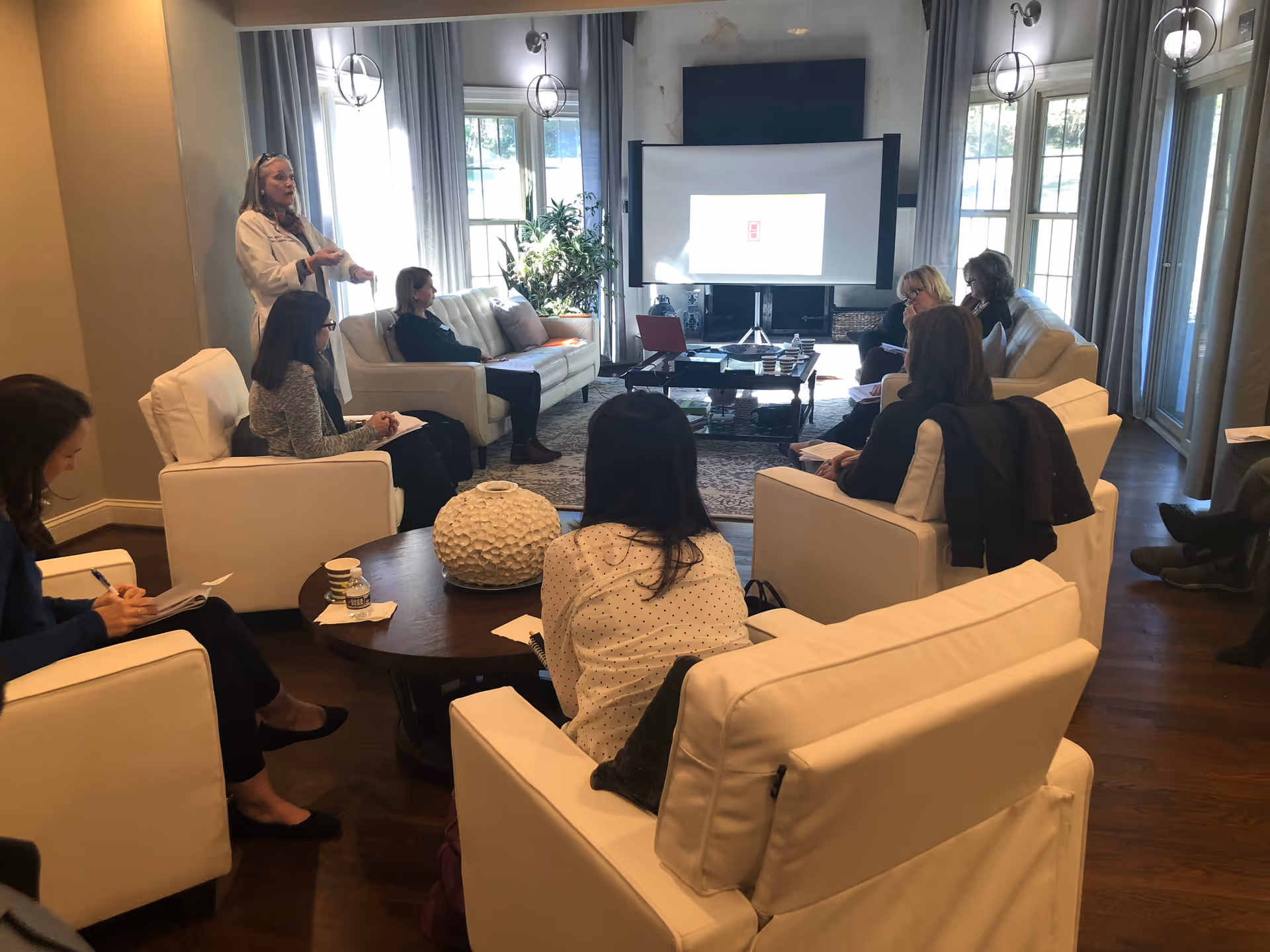A group of people seated in a bright living room listening to a presenter with a projection screen at the front.