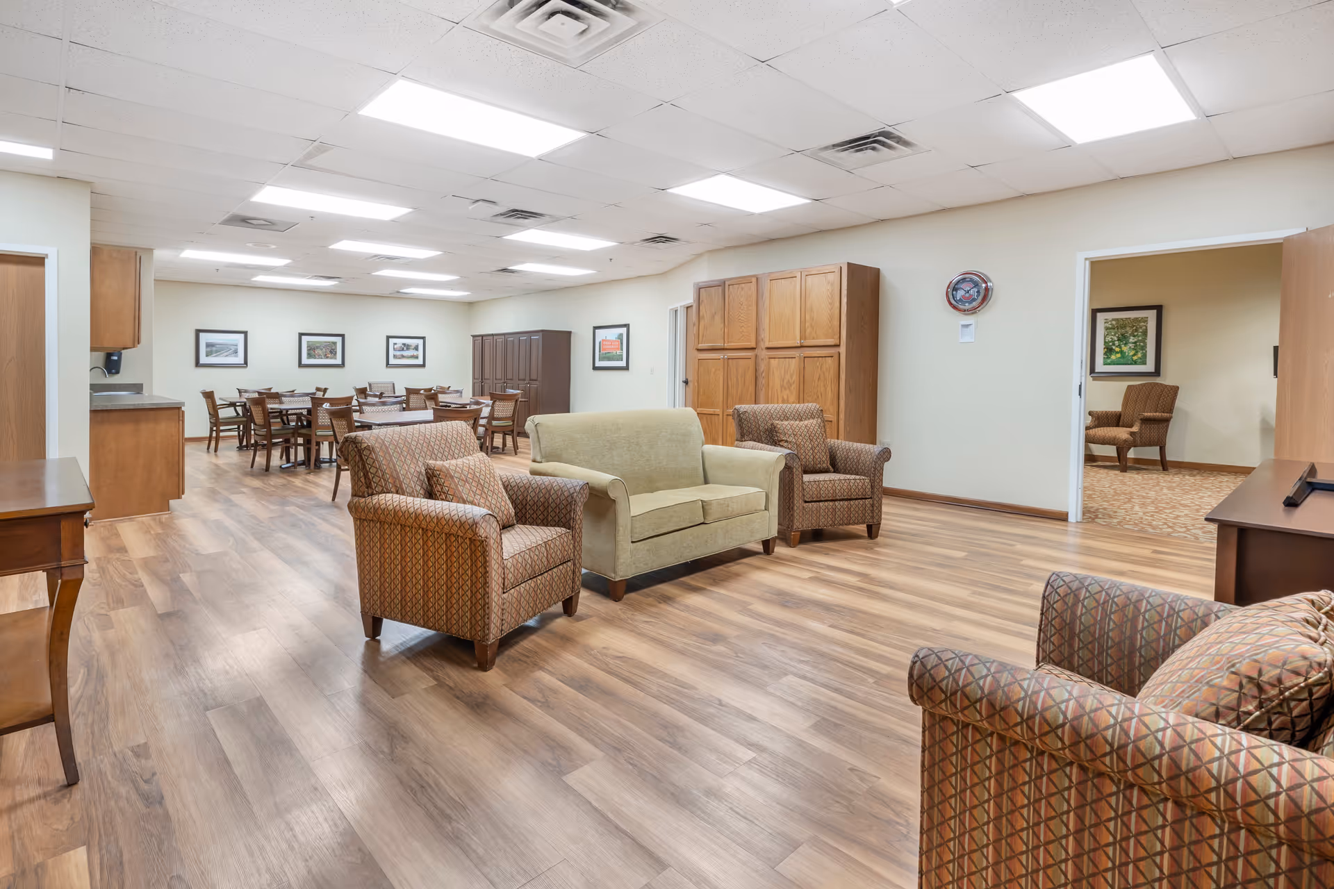 A spacious senior living facility common area with wood flooring, several upholstered armchairs and a loveseat arranged in the foreground, and multiple wooden dining tables with chairs in the background. The walls are light-colored with framed pictures and a clock. There are two open doorways leading to other rooms, and the ceiling has recessed lighting and air vents.