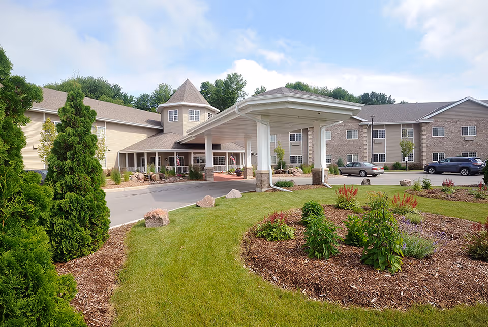 Exterior view of Emerald Bay Retirement Community featuring a covered entrance with white pillars, surrounded by well-maintained landscaping including green grass, shrubs, and flowers. The building has beige and brown siding with multiple windows and a turret-like structure.