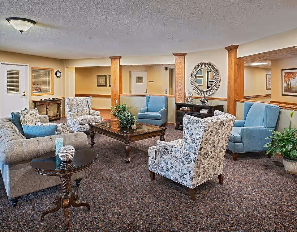 A cozy senior living facility common area with a mix of upholstered armchairs and a sofa arranged around a wooden coffee table. The room features warm beige walls, wooden columns, a round decorative mirror, framed artwork, and potted plants, creating a welcoming and comfortable atmosphere.