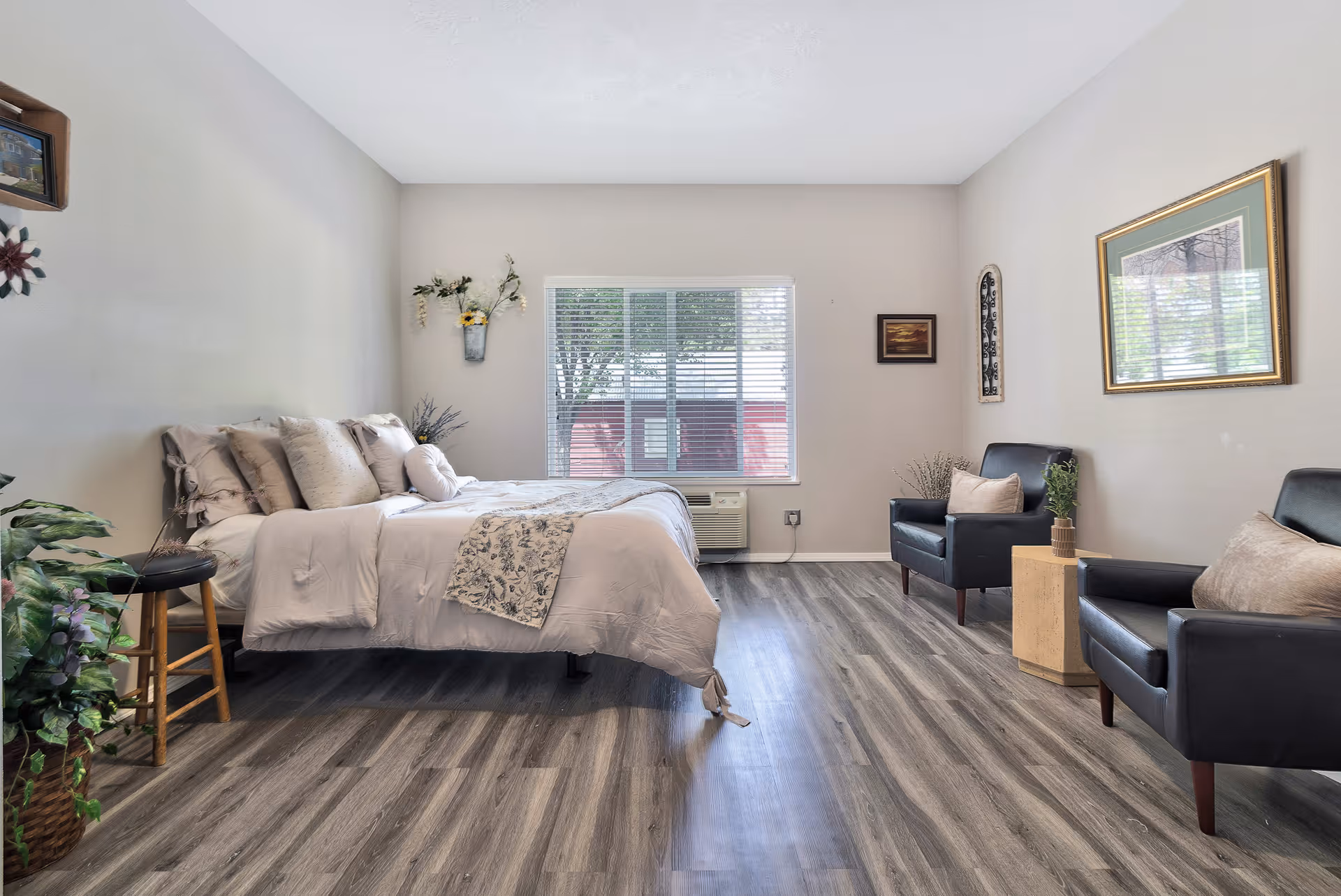 A bright and spacious senior living bedroom with a neatly made bed featuring multiple pillows and a floral throw. The room has wood-style flooring, two black armchairs with beige cushions, a small wooden side table with a plant, and a large window with blinds letting in natural light. The walls are decorated with framed pictures and a small wall-mounted flower arrangement.
