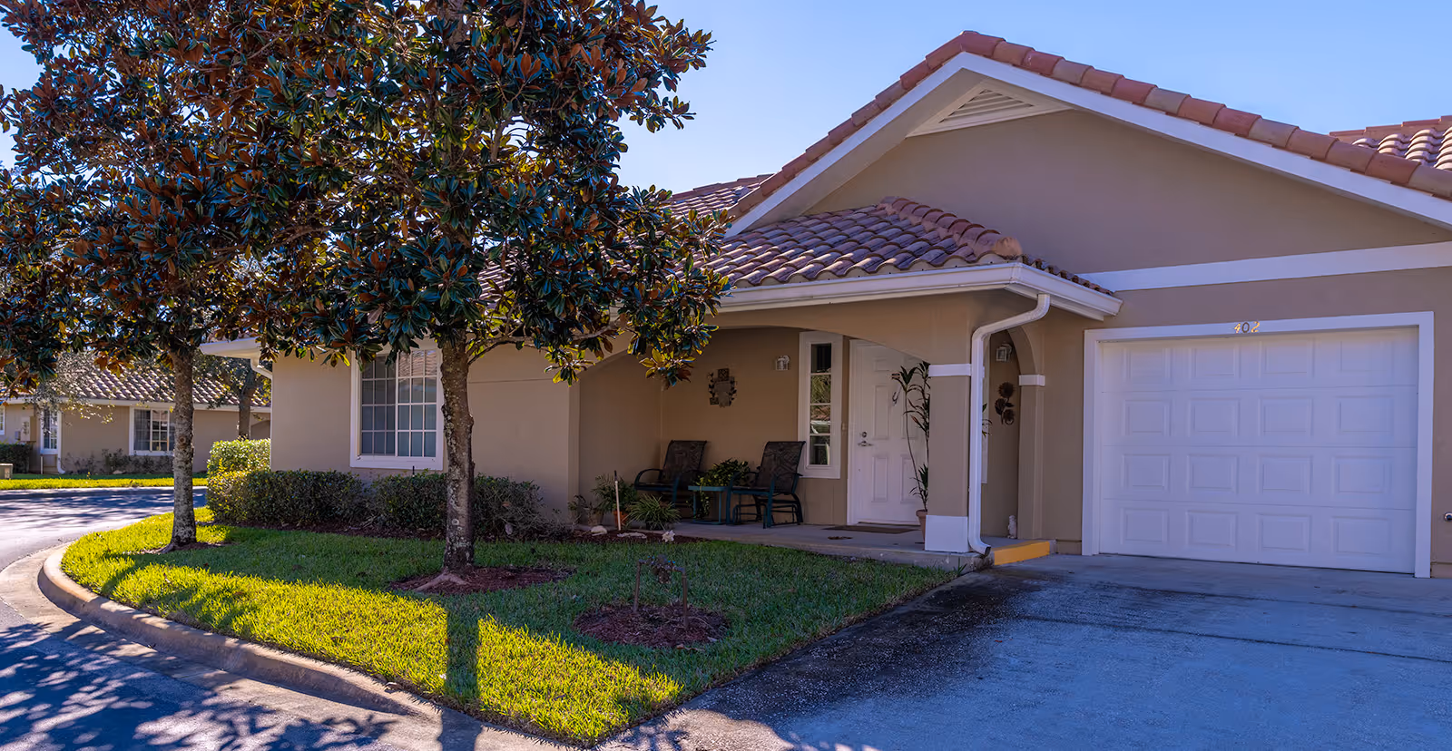 Exterior view of a single-story residential unit at Sterling Court - Sky Active Living, featuring a beige stucco facade, a tiled roof, a white garage door, a small covered porch with two chairs and a table, and a well-maintained lawn with two trees in front.