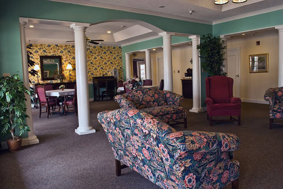 Carpeted common room with floral-upholstered sofas and armchairs, decorative white columns, and a dining table area in the background.
