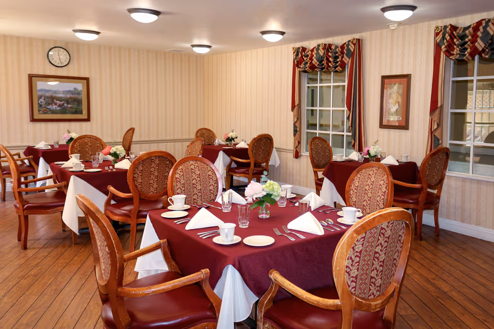 Dining room with multiple tables set with burgundy tablecloths, floral centerpieces, place settings, and upholstered wooden chairs.