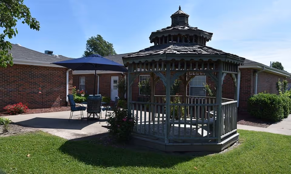 A wooden gazebo and patio with a table, umbrella, and chairs in a grassy courtyard outside a brick assisted-living building.