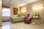 A nursing center room with a single bed covered in a green blanket, a purple recliner chair with a beige throw draped over it, a small round wooden table with a floral arrangement, a window with blinds and a valance, a wall-mounted air conditioning unit, and soft lighting fixtures on the wall.