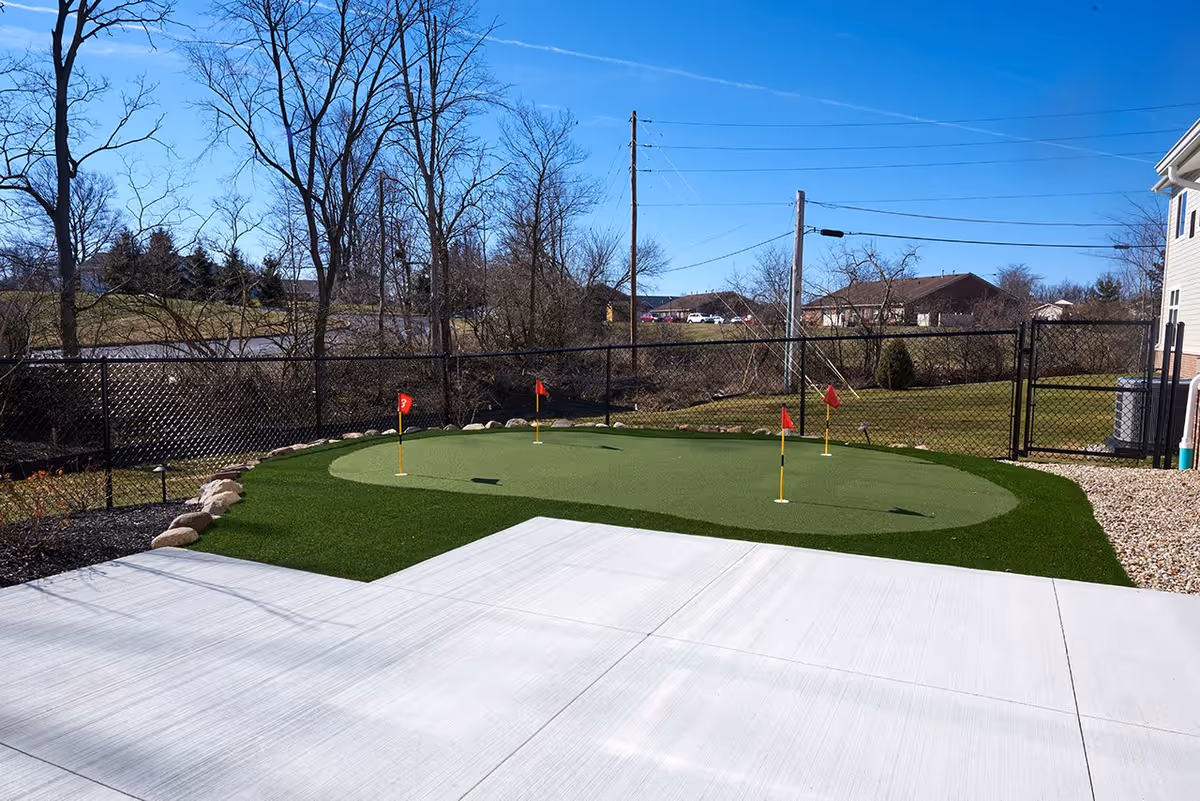 Outdoor area with a small putting green featuring five red flags on poles, surrounded by a black chain-link fence. There is a concrete patio in the foreground and leafless trees and buildings in the background under a clear blue sky.