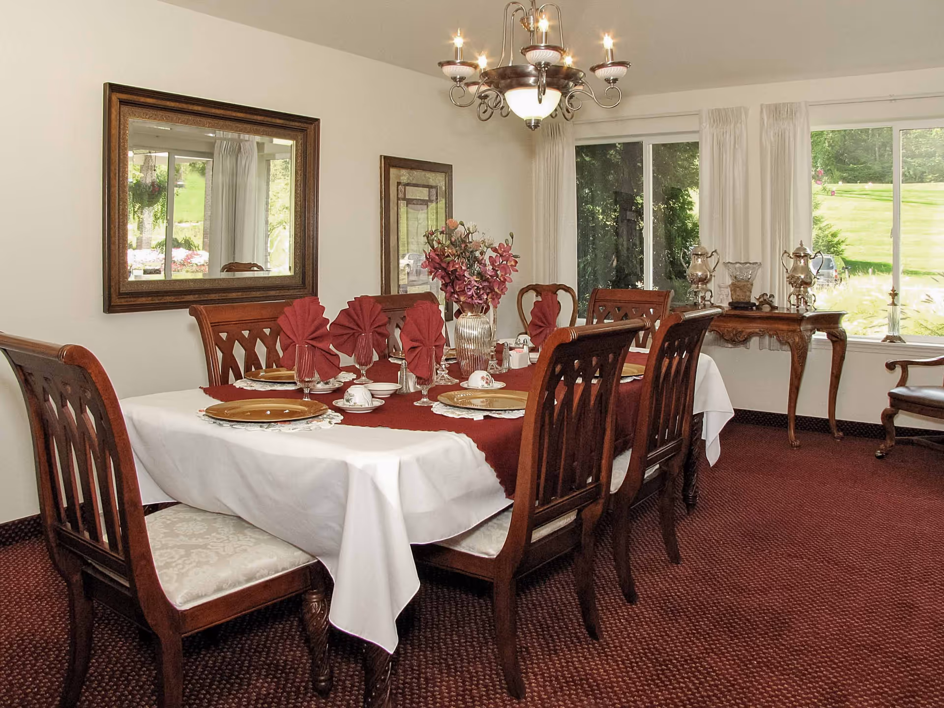 Formal dining room with a table set for a meal, wooden chairs, a chandelier, and large windows overlooking a lawn.