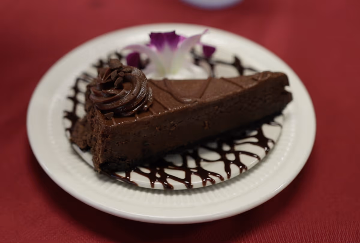 A slice of chocolate cake with chocolate frosting and chocolate drizzle on a white plate, garnished with a purple and white flower, placed on a red tablecloth.