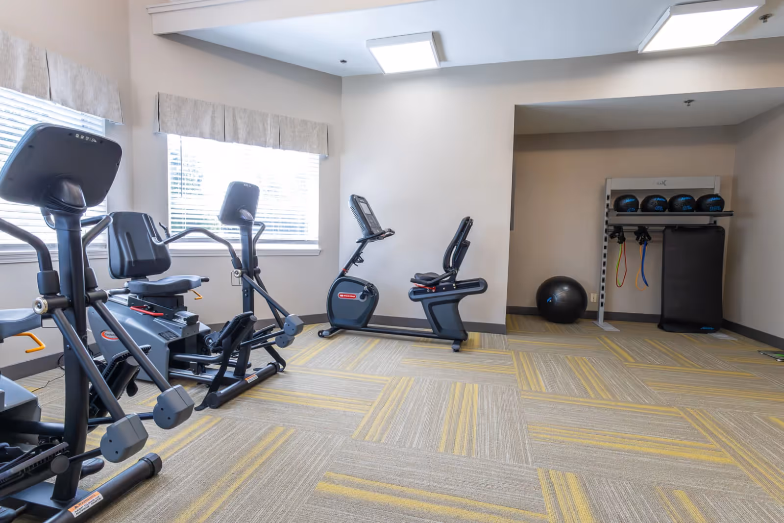 A small fitness room with exercise equipment including two recumbent stationary bikes, an elliptical machine, and a rack holding medicine balls, resistance bands, and a large exercise ball. The room has light-colored walls, a window with blinds and a valance, and patterned carpet with yellow and gray tones.