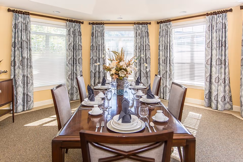 A bright dining room with a polished wooden table set for six people. Each place setting includes a white plate, bowl, silverware, a wine glass, and a black folded napkin. The room has beige walls and large windows with patterned curtains allowing natural light to fill the space. A floral centerpiece decorates the middle of the table.