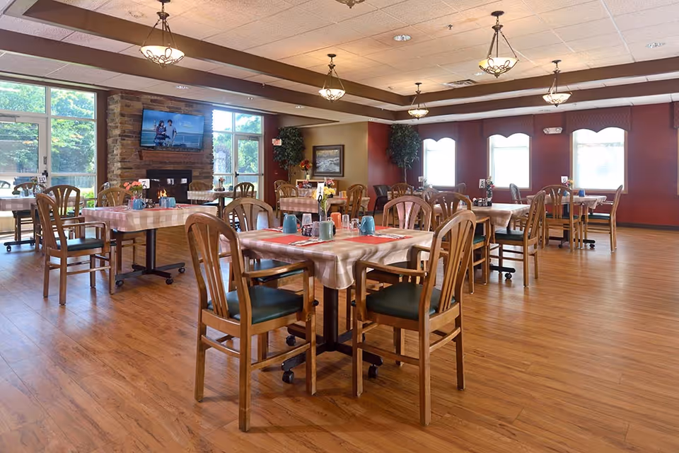 A spacious dining room with wooden floors and multiple tables covered with checkered tablecloths. Each table has wooden chairs with green cushions and is set with cups, napkins, and condiments. Large windows let in natural light, and a stone fireplace with a mounted TV is visible on one wall. The room has warm lighting from ceiling fixtures and decorative plants near the walls.