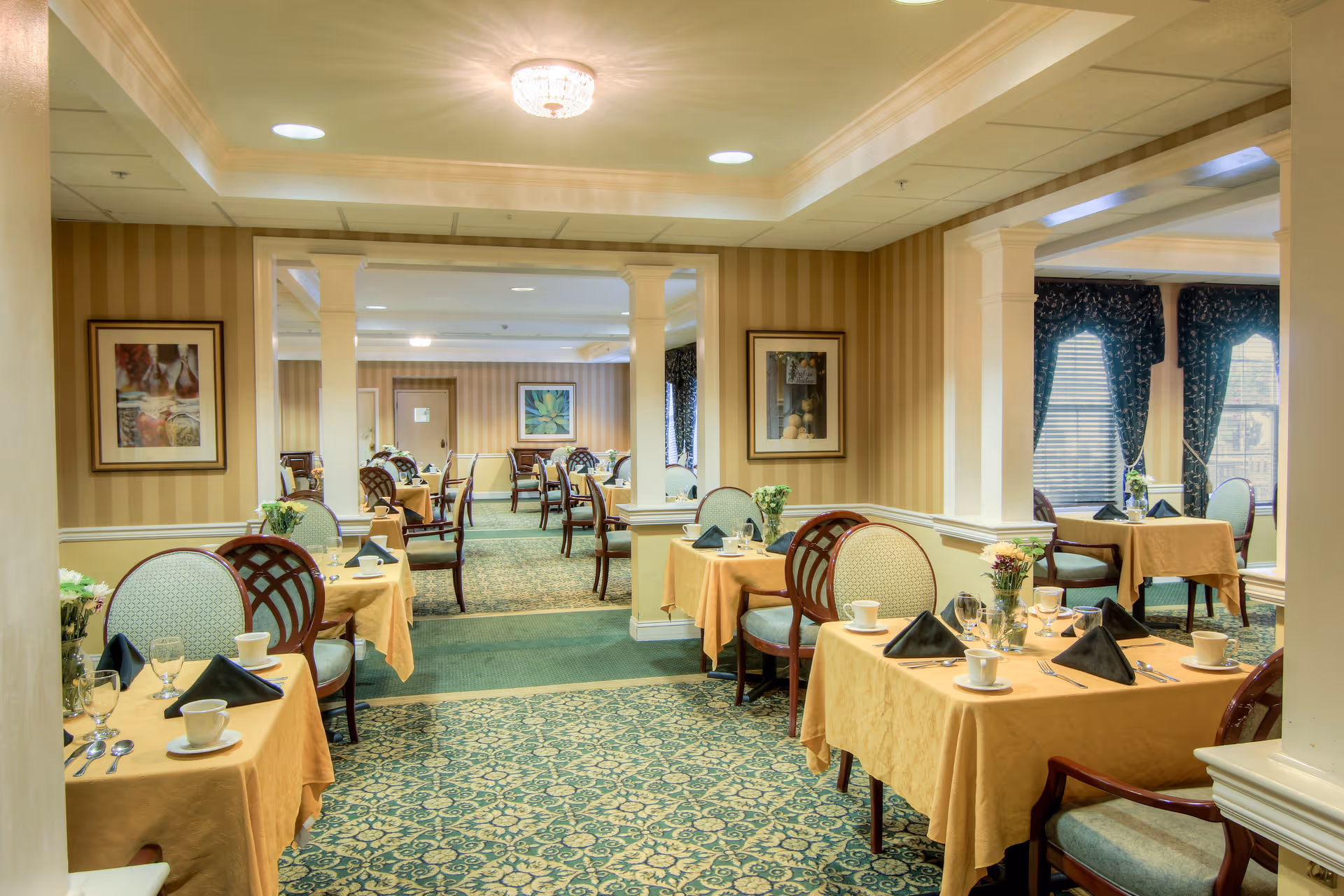 A well-lit dining room in a senior living facility with multiple tables covered in yellow tablecloths, each set with cups, glasses, silverware, and black folded napkins. The room features patterned carpet, striped wallpaper, framed artwork on the walls, and large windows with dark curtains allowing natural light to enter.