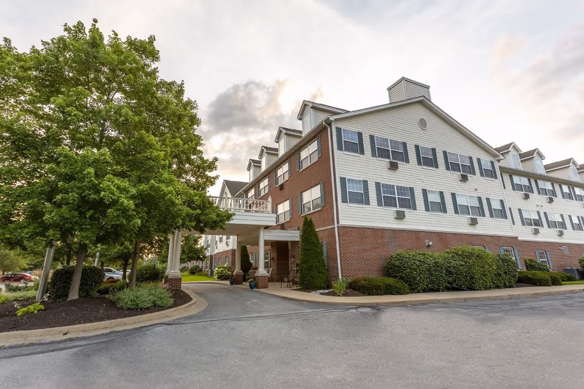Exterior view of a multi-story senior living facility building with brick and white siding, multiple windows with black shutters, and a covered entrance. There are green trees and shrubs around the building, and a paved driveway leading to the entrance.