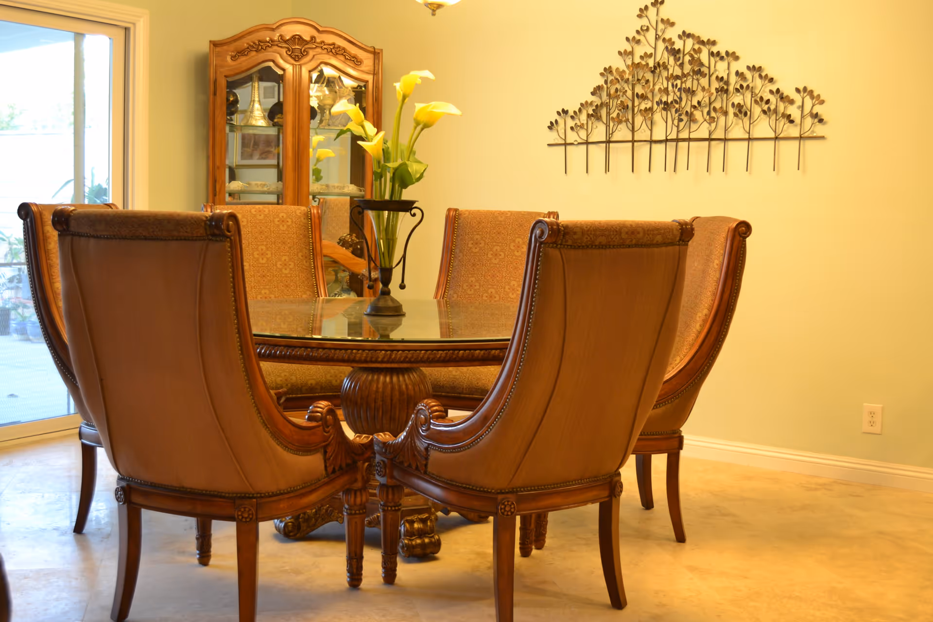 A dining room with a round glass-top table surrounded by six upholstered wooden chairs. A vase with yellow calla lilies is placed in the center of the table. Behind the table is a wooden china cabinet with glass doors displaying decorative items. A metal wall art piece with a tree branch design is mounted on the light-colored wall. A sliding glass door is visible on the left side, allowing natural light into the room.