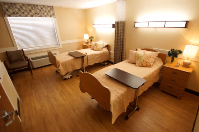 A nursing home bedroom with two single beds, each with beige bedspreads and decorative pillows. Each bed has a small overbed table. There are two wooden nightstands with lamps and flowers, a window with blinds and a patterned valance, and two chairs near the window. The room has wood flooring and soft yellow walls with modern wall-mounted lights above each bed.