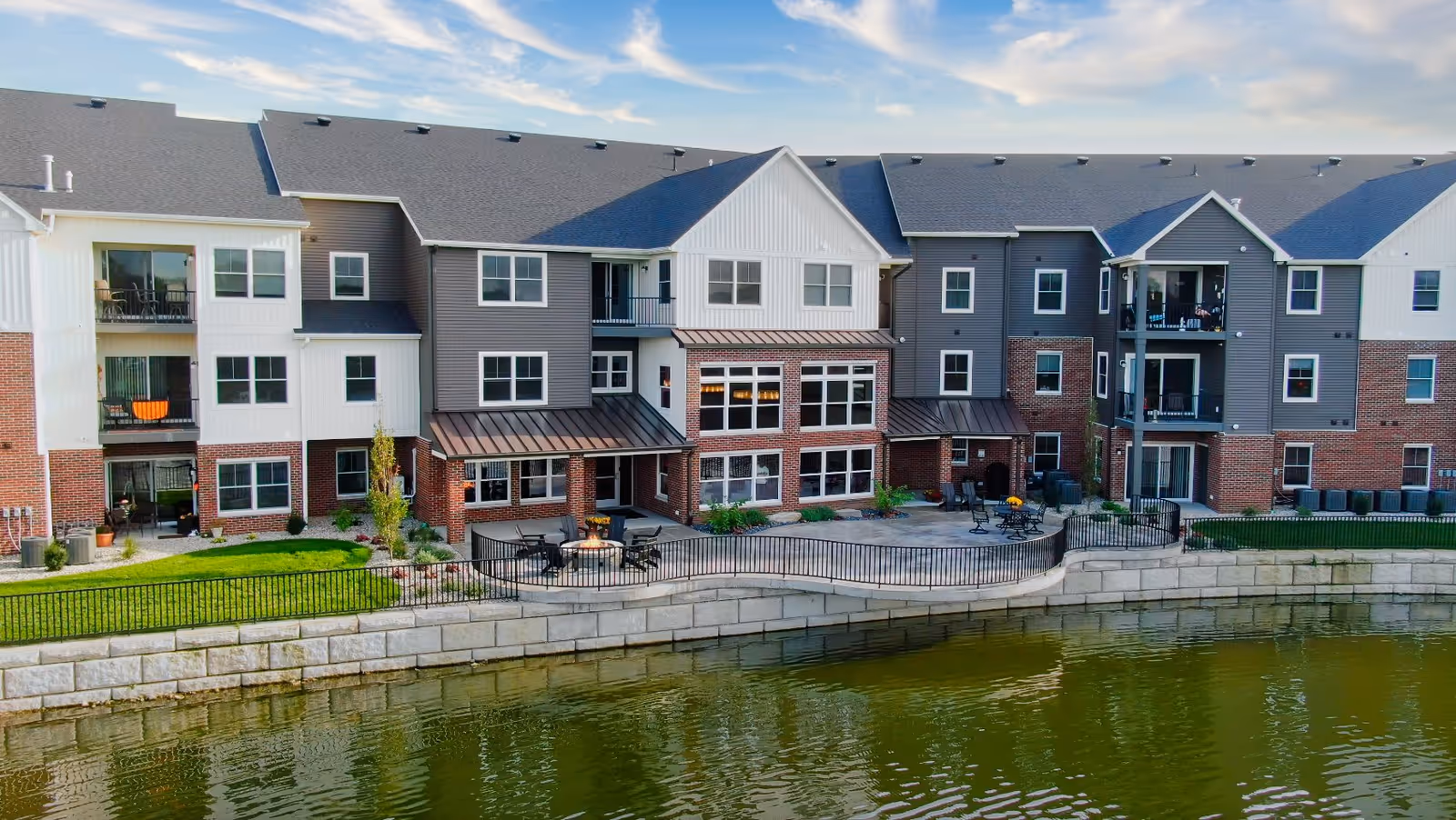 Exterior view of a multi-story senior living facility building with balconies, large windows, and a patio area with outdoor seating and a fire pit, situated next to a body of water with a stone retaining wall.