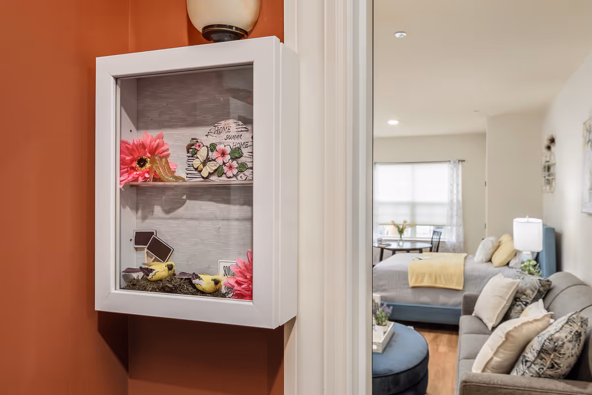 A decorative white shadow box mounted on a burnt orange wall containing pink flowers, small yellow bird figurines, and a sign that reads 'Home Sweet Home'. In the background, a cozy bedroom is visible with a bed, a small dining table by a window, and a gray sofa with pillows.