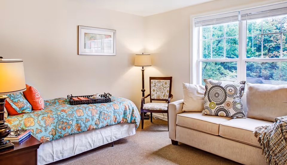 Sunlit furnished bedroom with a patterned bed, beige sofa, armchair, floor lamp, and a large window showing greenery.