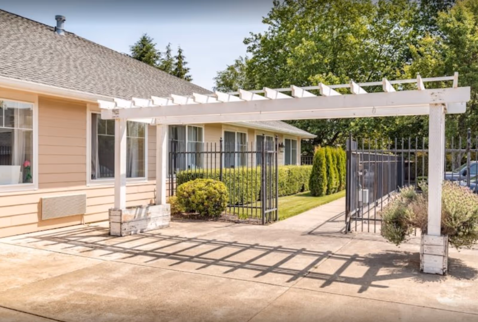 Outdoor view of a senior living facility with a beige building, large windows, a white wooden pergola casting shadows on the concrete ground, black metal fencing, green bushes, and trees in the background under a clear sky.