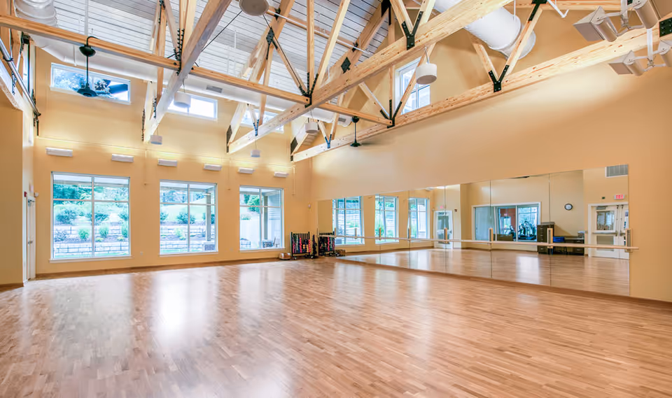 Spacious room with wooden floor and large windows allowing natural light, featuring high ceilings with exposed wooden beams and ceiling fans. One wall is covered with large mirrors and ballet barres, suggesting a fitness or dance studio environment.