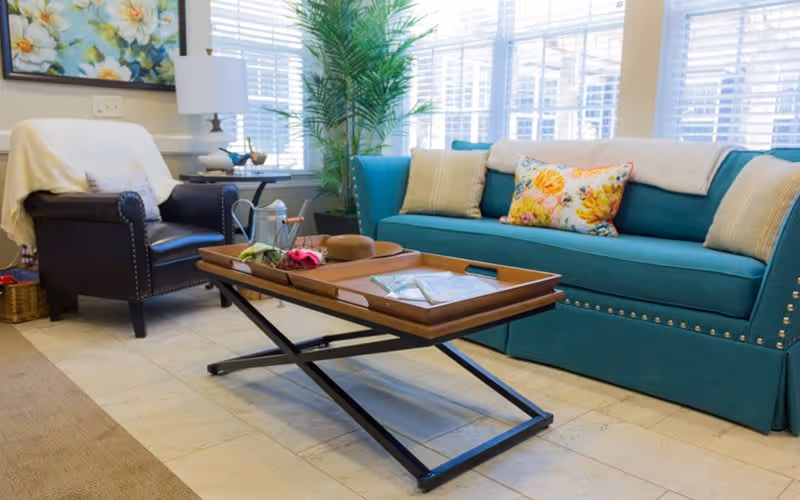 A cozy living room with a teal sofa adorned with floral and solid pillows, a dark leather armchair with a white throw draped over it, a wooden coffee table with a metal frame holding a tray with decorative items, a potted plant, a side table with a lamp, and large windows with blinds letting in natural light.