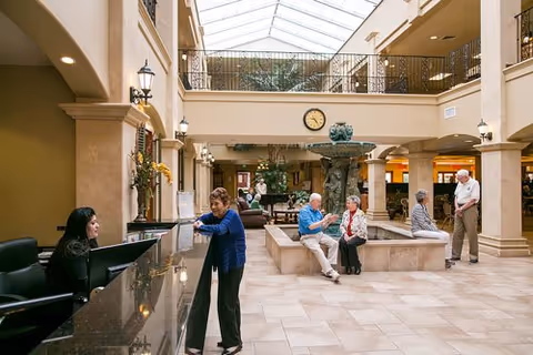 A spacious and well-lit lobby area with a high glass ceiling. Several elderly people are seen interacting and sitting around a central water fountain. A woman is standing at a reception desk talking to a receptionist. The area has beige tiled floors, columns, and a clock on the wall above the fountain.