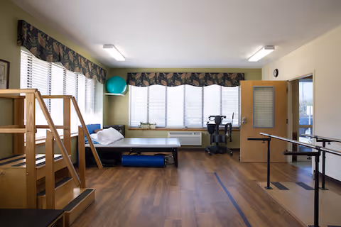 A spacious therapy/exercise room with wooden floors, parallel bars, a treatment table, exercise equipment and large windows with blinds and patterned valances.