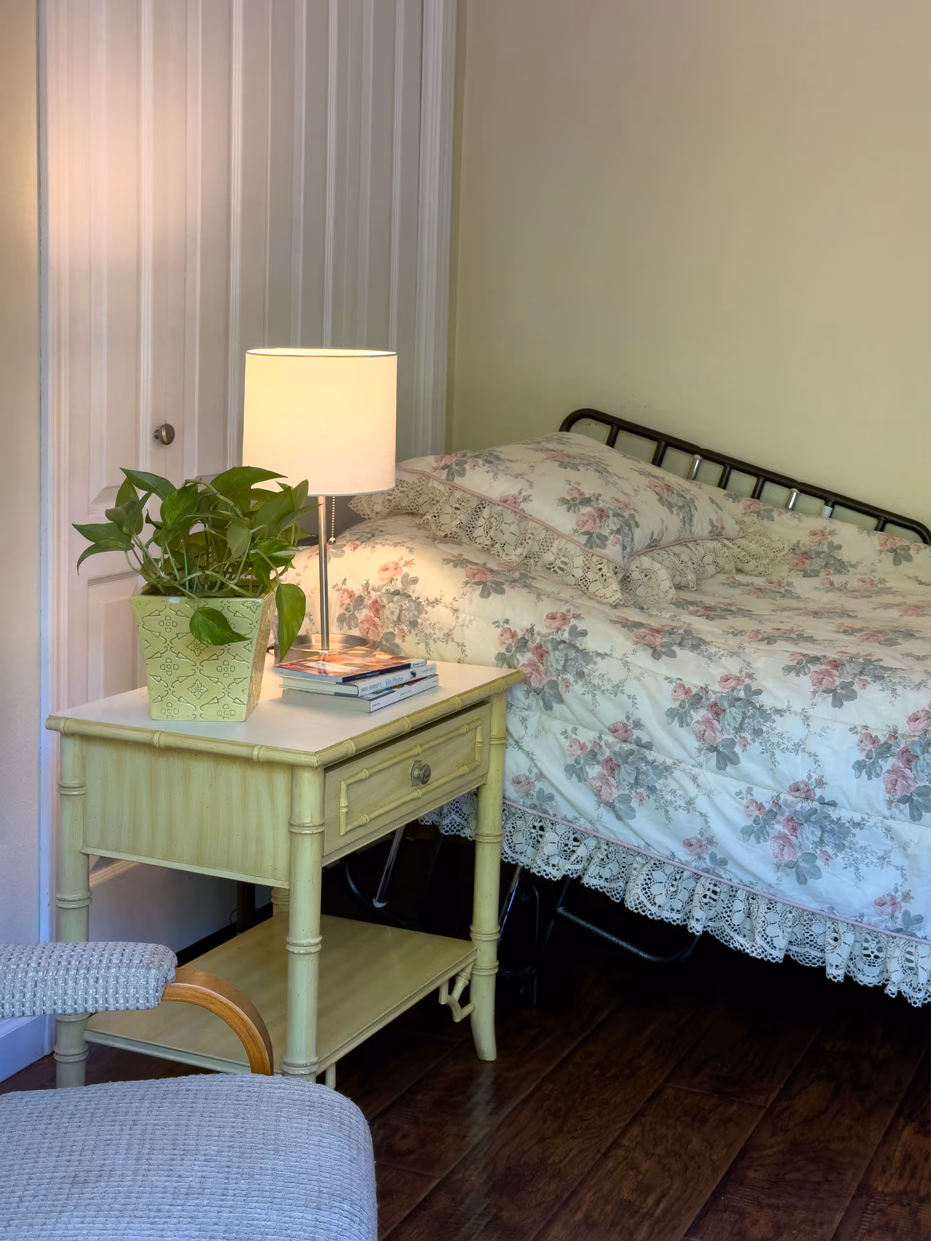 A cozy bedroom corner featuring a bed with floral-patterned bedding and lace trim, a light wooden nightstand with a potted plant, a lamp, and a few books on top. A cushioned chair is partially visible in the foreground, and a white paneled door is in the background.