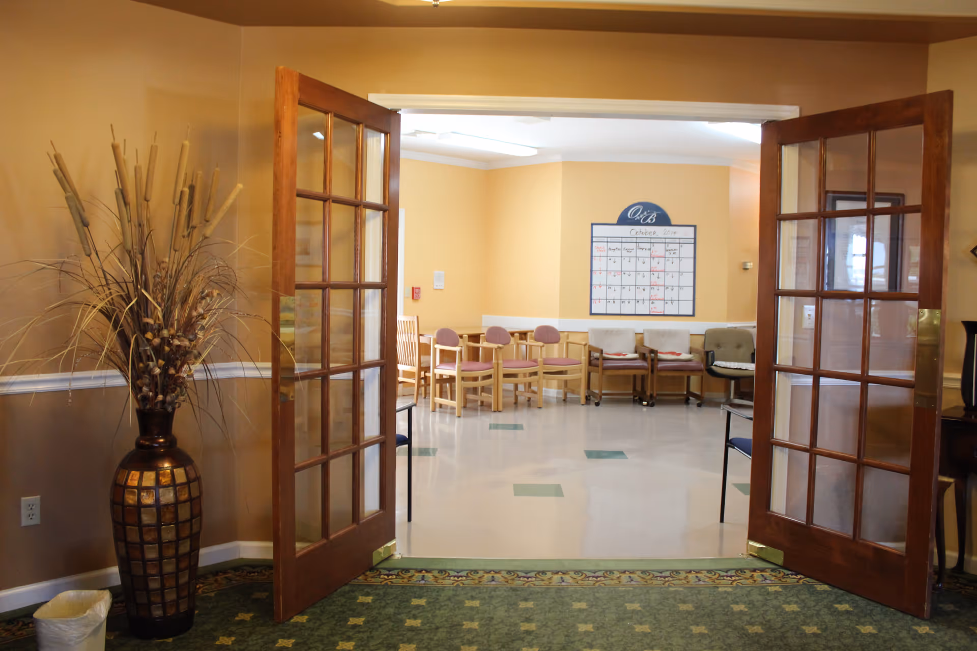 View through open double wooden doors with glass panes into a room with chairs arranged along the walls and a large calendar on the far wall. A tall decorative vase with dried plants is positioned to the left of the doors, and the floor has a green patterned carpet transitioning to a tiled floor inside the room.
