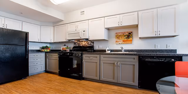 Modern kitchen with white upper cabinets and gray lower cabinets, black appliances including refrigerator, stove, microwave, and dishwasher, a wooden floor, and a small round glass table with a red chair.