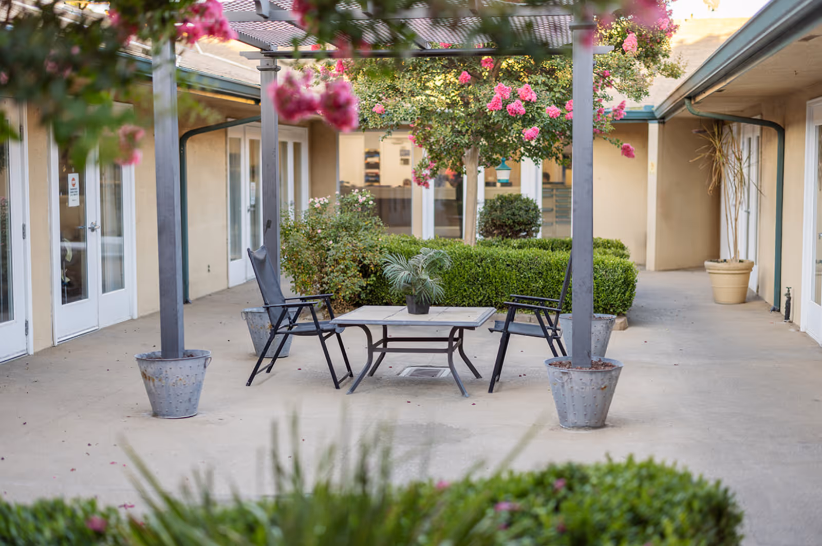 Outdoor courtyard with a pergola, table and chairs, potted plants and flowering bushes between building entrances.