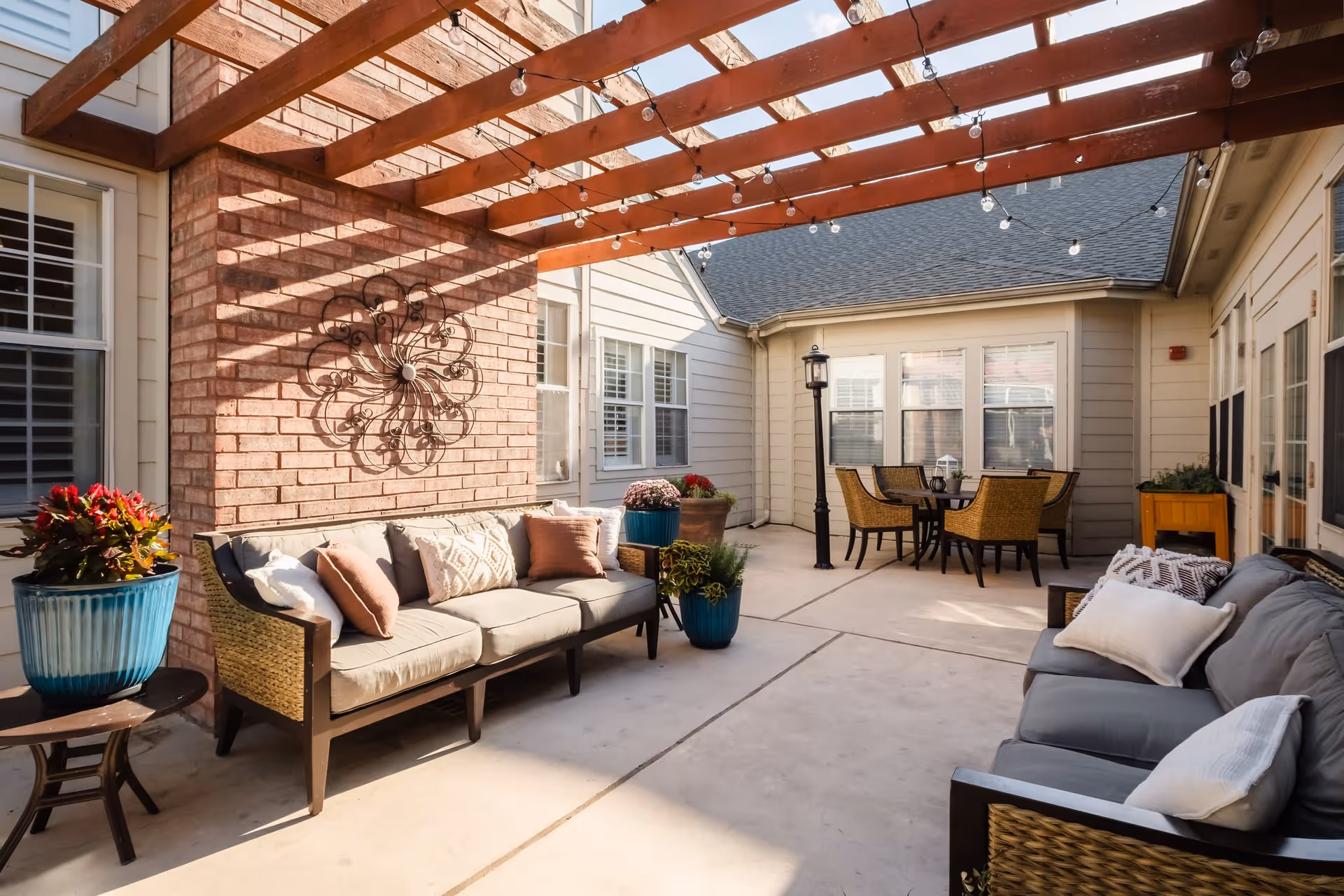 Outdoor patio area with two cushioned sofas, decorative pillows, potted plants, a wooden pergola with string lights overhead, and a round table with four wicker chairs near the building windows.