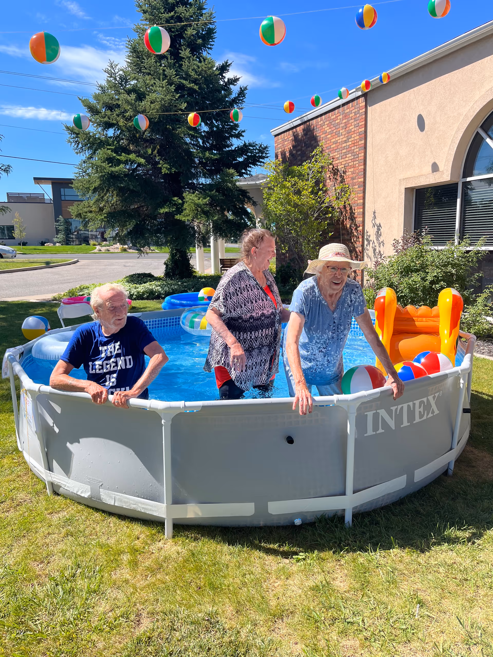 Three elderly people enjoying a sunny day in a small above-ground pool in a grassy outdoor area. Colorful beach balls are floating in the pool and hanging on strings above. One person is wearing a hat and smiling, another is standing in the pool, and the third is sitting on the edge. The background shows a building with a large window and some greenery.