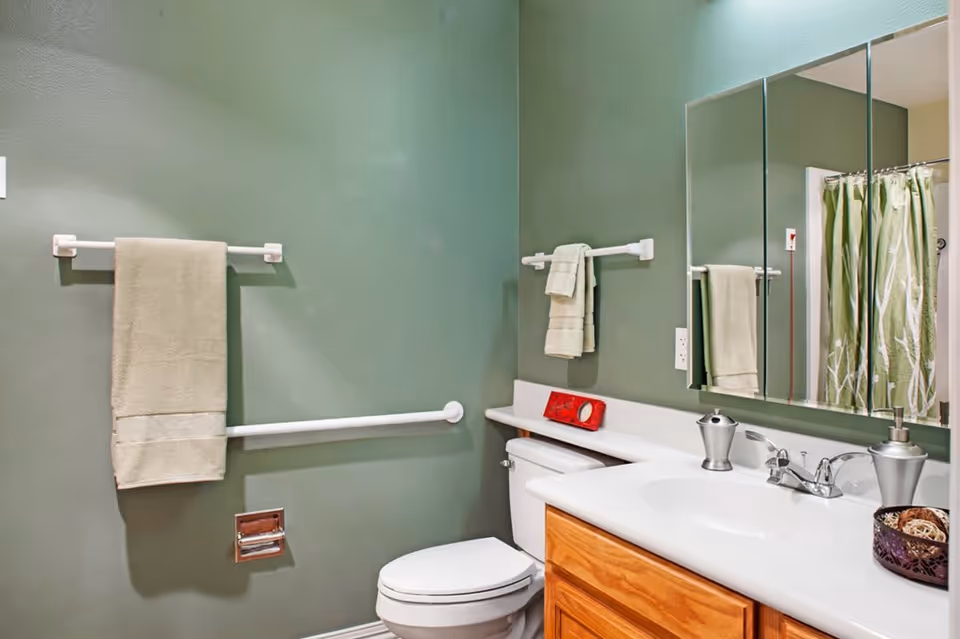 A bathroom with green walls featuring a white toilet, a wooden vanity with a white countertop and sink, a large mirror above the sink, towel racks with beige towels, and a shower with a green and white patterned curtain.