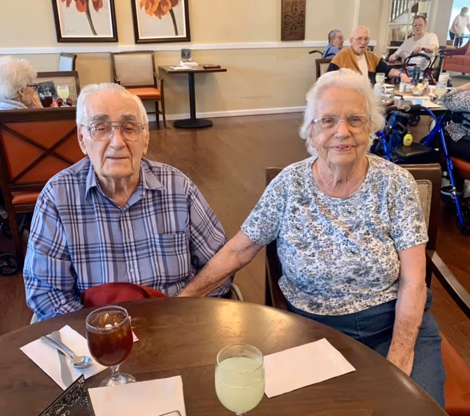An elderly man and woman seated at a dining table in a senior living dining room, smiling and holding hands.