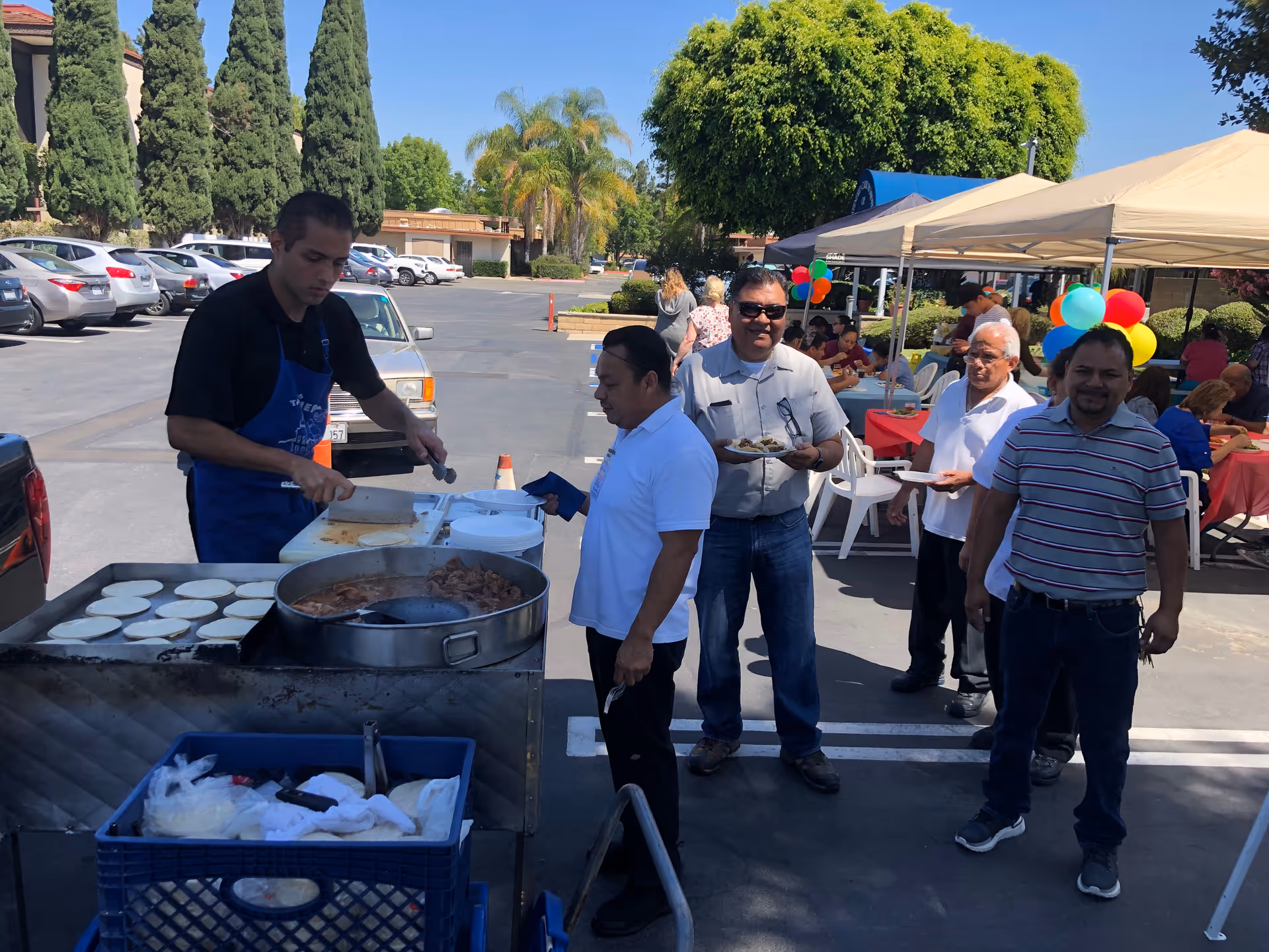 Outdoor event at Extended Care Hospital of Westminster with a man cooking food on a large grill while several people wait in line holding plates. There are tents with tables and chairs in the background where people are seated, colorful balloons, and trees surrounding the parking lot area.