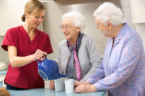 A caregiver pours tea from a blue teapot into mugs while two older women watch and smile in a kitchen setting.