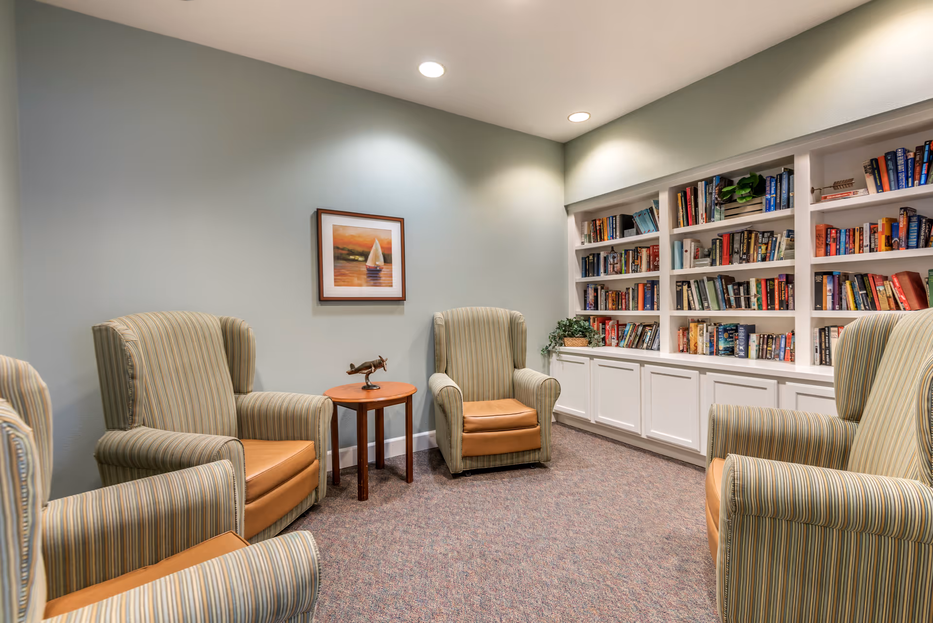 A cozy reading or sitting area with four striped armchairs arranged around a small wooden table. The room has a built-in white bookshelf filled with books and a framed picture of a sailboat on the wall. The lighting is soft with recessed ceiling lights.